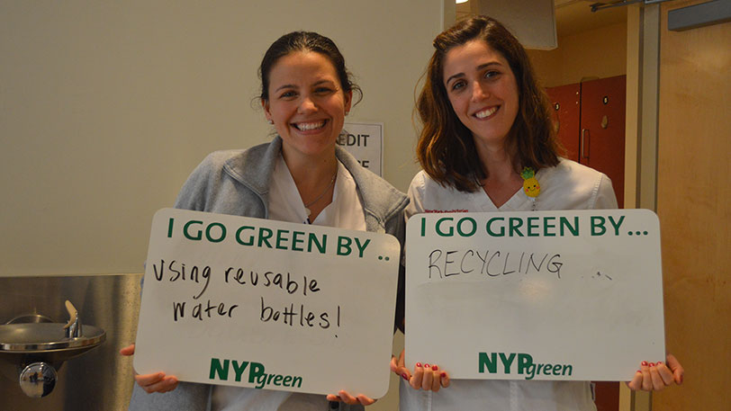 Two women holding signs that say I go green by... using reusable water bottles and I go green by... recycling