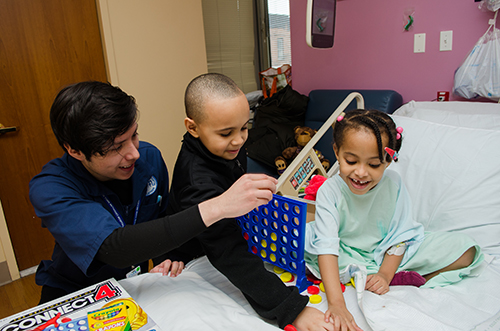 Child playing connect 4 in a hospital bed with sibling and nurse
