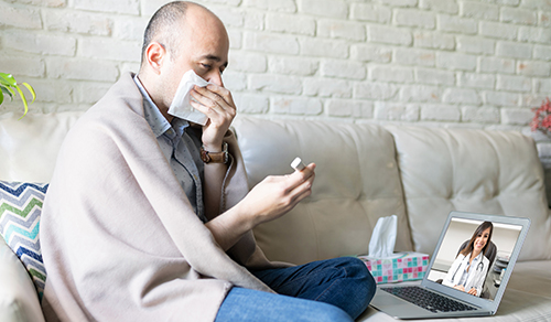 image of man covering mouth holding thermometer in front of doctor on laptop