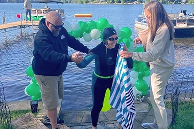 Bonnie Dragland wearing a wetsuit, swim cap, and goggles is helped out of a lake by two supporters—one man and one woman—on a sunny day. Green and white balloons form an arch behind them near a dock with boats, suggesting the end of a swimming event or challenge